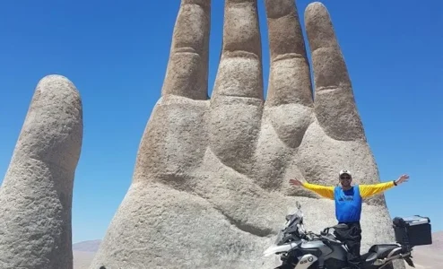 Homem em moto frente a escultura mão gigante deserto.