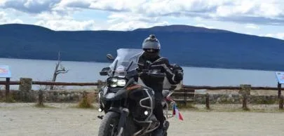 Motociclista em estrada de terra com lago e montanhas.