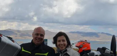 Casal sorridente com capacetes, montanhas ao fundo, céu nublado.