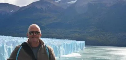 Homem sorrindo em frente a geleira e montanhas ao fundo.