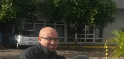 Homem sorrindo em frente a prédio com árvores ao fundo.
