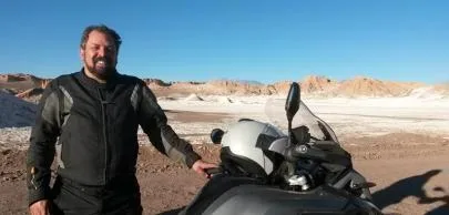 eb1d7003c8e6df756d2d2496e82e364b482d70a1 Homem sorridente ao lado de moto em paisagem desértica.