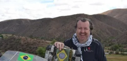 Homem sorridente com moto decorada em paisagem montanhosa.