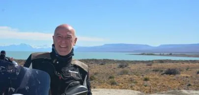 Homem sorrindo em paisagem de lago e montanhas ao fundo.