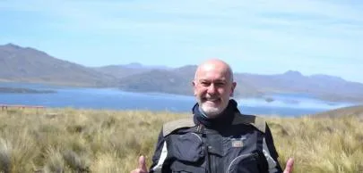 Homem sorridente em campo aberto com lago ao fundo.