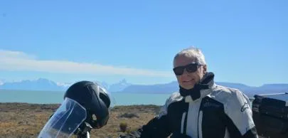 Homem de óculos ao lado de moto em paisagem montanhosa.