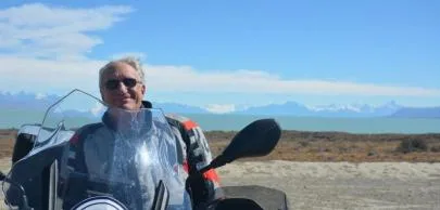 Homem sorrindo em motocicleta com paisagem montanhosa ao fundo.