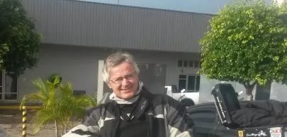 Homem sorridente em frente a prédio, árvores ao fundo.