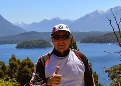 Homem sorrindo com paisagem de lago e montanhas ao fundo.