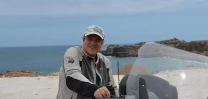 eb1d7003c8e6df756d2d2496e82e364b482d70a1 Homem sorridente em motocicleta à beira-mar ensolarada.