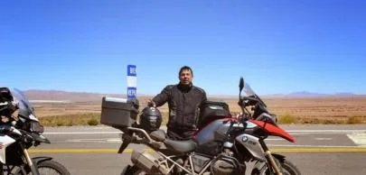 Homem com motocicleta em estrada deserta sob céu azul.