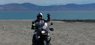 Motociclista acena em estrada deserta com lago ao fundo.