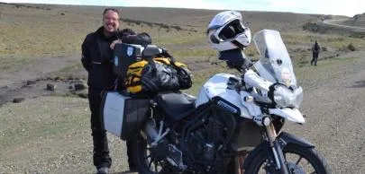 Homem sorridente ao lado de moto carregada em estrada deserta.