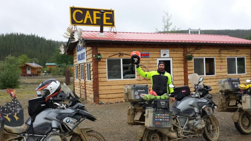 Piloto ao lado de sua moto que está parada na frente de um café 