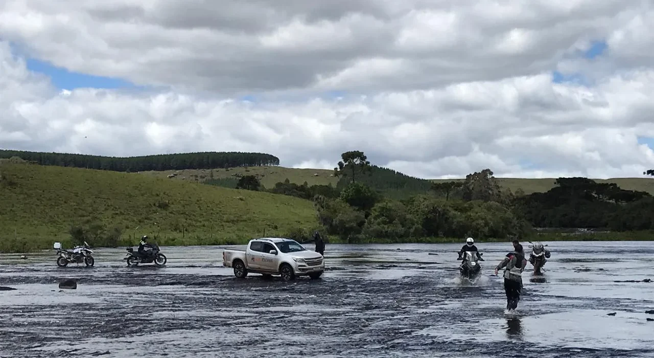 Pessoas e veículos atravessando rio raso em dia nublado.