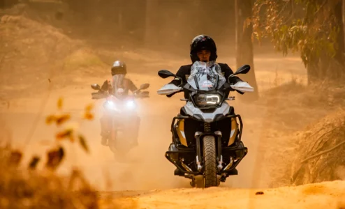 Dois motociclistas em estrada de terra com poeira levantada.