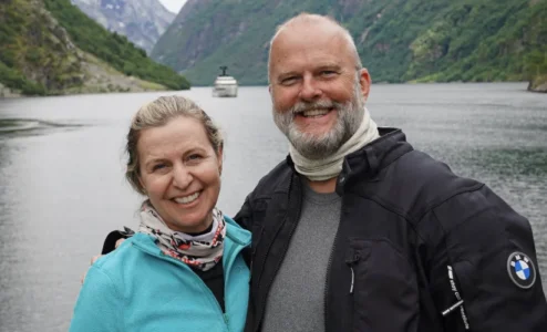 Casal sorridente em frente a fiorde com barco ao fundo.