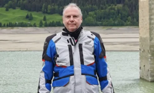 Homem em traje de motociclista em frente a lago alpino.