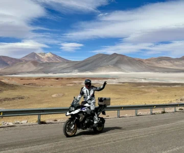 eb1d7003c8e6df756d2d2496e82e364b482d70a1 Motociclista acena em estrada deserta com montanhas ao fundo.